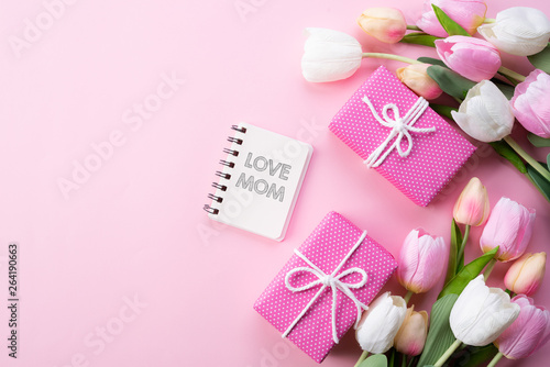 Happy mothers day concept. Top view of pink tulip flowers, gift box and note book with LOVE MOM text on pink pastel background. Flat lay.
