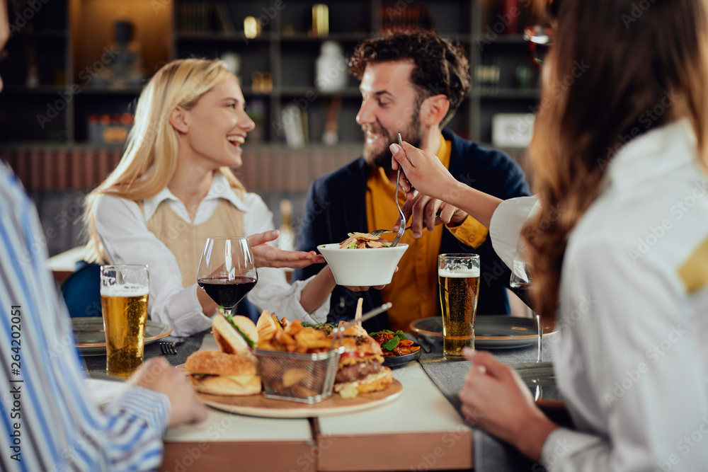 Multiethnic friends sitting at restaurant, drinking alcohol, chatting and having burgers for dinner.