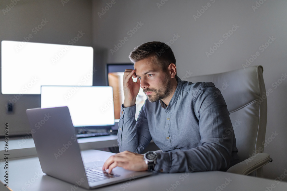 Young Caucasian serious handsome bearded employee using laptop for important project while sitting in office late at night. Young Caucasian serious handsome bearded employee using laptop for important project while sitting in office late at night.