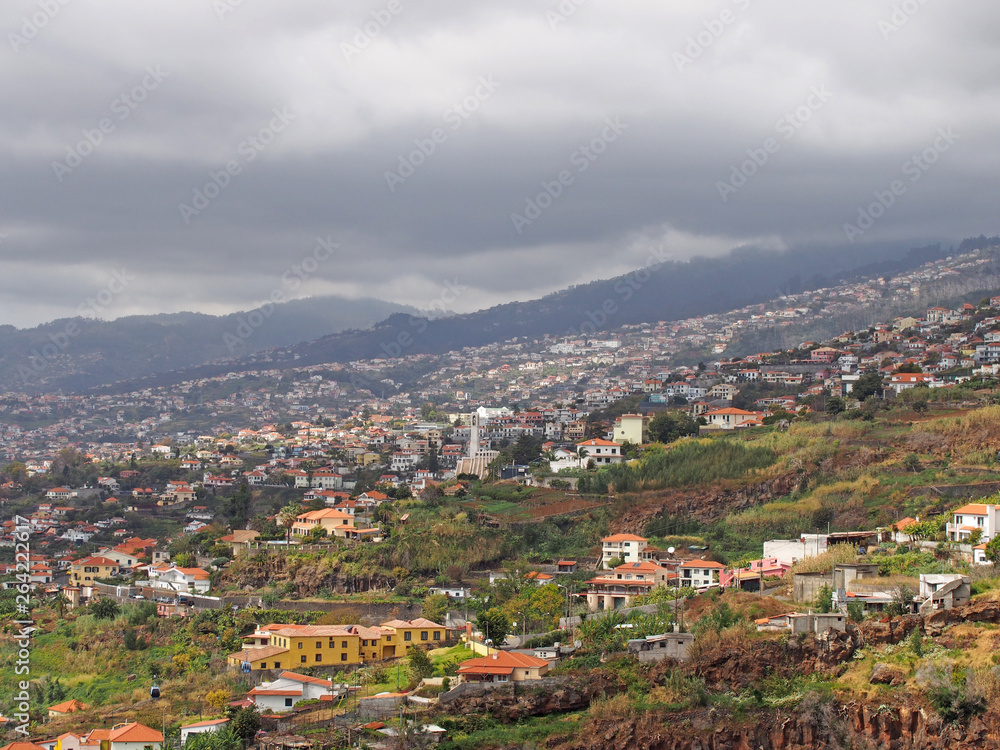a panoramic view of funchal in madeira showing small farms and agriculture with buildings of the city against distant cloudy mountains