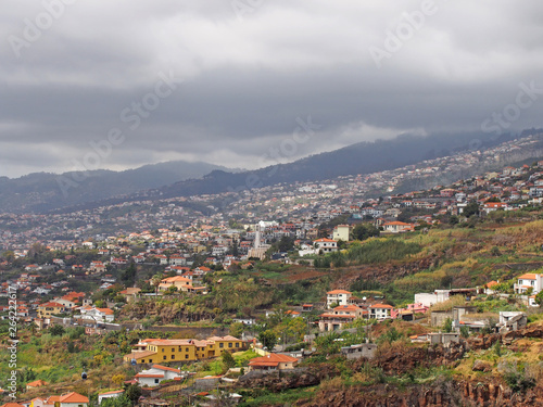 a panoramic view of funchal in madeira showing small farms and agriculture with buildings of the city against distant cloudy mountains