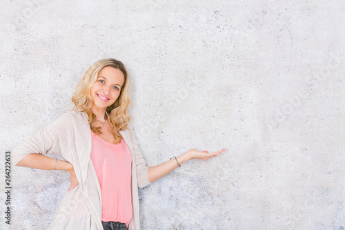 beautiful, blond woman is pointing at and posing in front of grey, concrete wall background