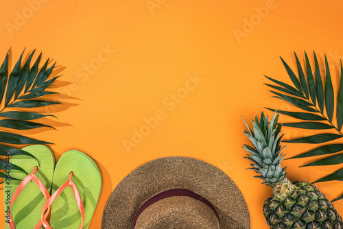top view of pineapple, tropical leaves, green flip flops and brown straw hat on orange background with copy space