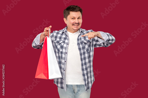 Cheerful modern middle aged man in white t-shirt and checkered shirt standing and pointing finger to shopping bags and toothy smile, looking at camera. indoor studio shot on isolated red background.