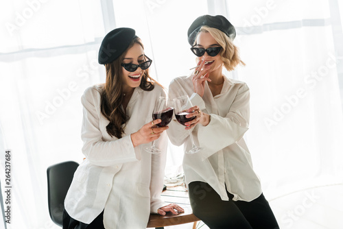 elegant happy brunette and blonde women in black berets and sunglasses laughing, smoking and drinking red wine