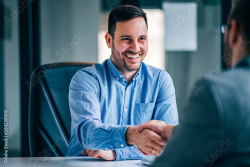 Two smiling businessmen shaking hands while sitting at the office desk.