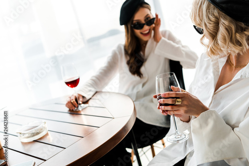 selective focus of blonde woman drinking wine with elegant smiling and surprised brunette friend in black beret and sunglasses