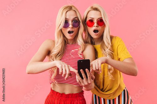 Shocked blonde twins in sunglasses using smartphone photo