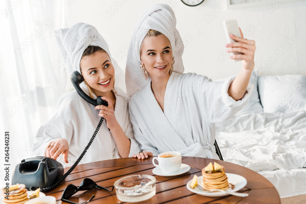 stylish women in bathrobes and jewelry with towels on heads talking on retro phone and smiling while taking selfie stylish women in bathrobes and jewelry with towels on heads talking on retro phone and smiling while taking selfie