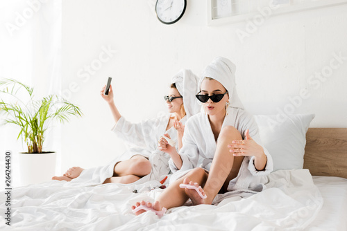 stylish women in bathrobes, sunglasses, towels and jewelry lying in bed, doing pedicure and taking selfie