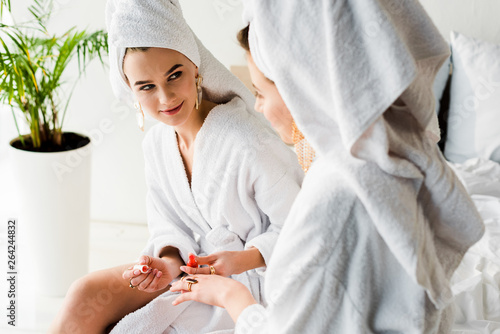 stylish woman in bathrobe and jewelry, with towel on head sitting on bed with nail polish and looking at friend