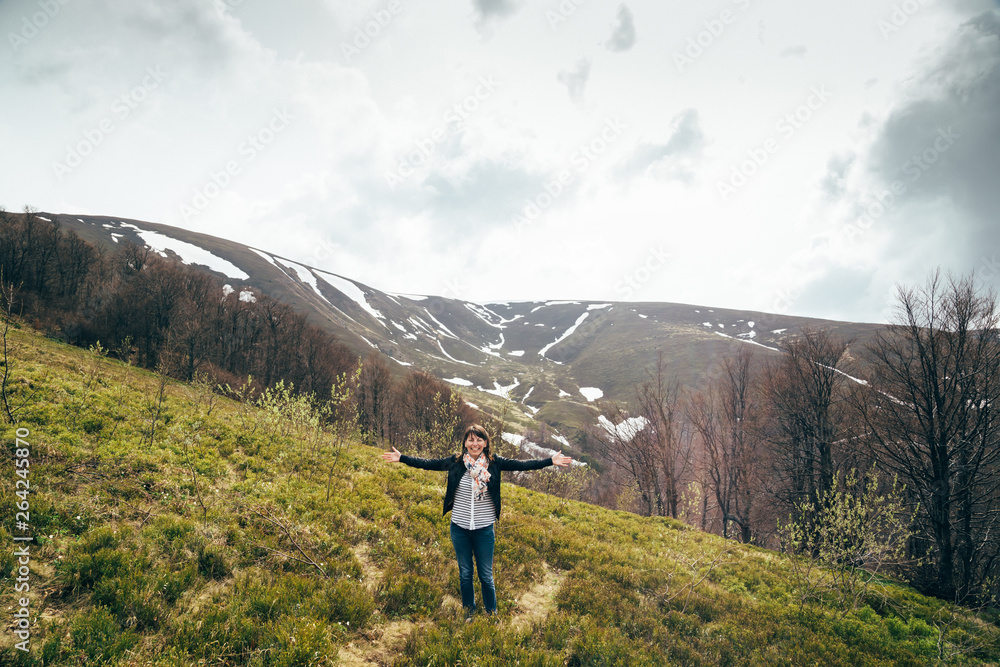 Happy smiling tourist girl in blue jeans and striped shirt in mountains surrounded by forest, raising arms and enjoying silence and harmony of nature