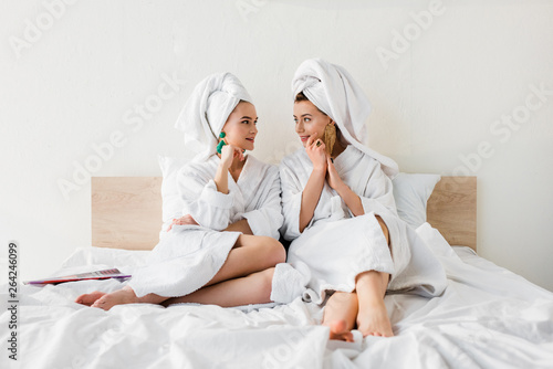 stylish girls in earrings, bathrobes and with towels on heads sitting on bed and looking at each other