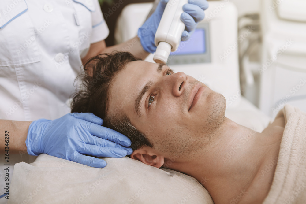 Close up of a handsome male client getting facial laser skincare treatment by professional dermatologist. Cosmetologist using cavitation laser apparatus on a male client. Rejuvenating concept Close up of a handsome male client getting facial laser skincare treatment by professional dermatologist. Cosmetologist using cavitation laser apparatus on a male client. Rejuvenating concept