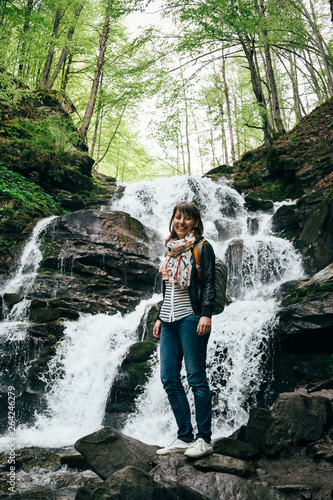 Happy smiling tourist girl in blue jeans  striped shirt and black jacket with backpack near to the cold fresh waterfall. Waterfall Shypit  cascade in Pylypets in forest. Carpathian Mountains  Ukraine