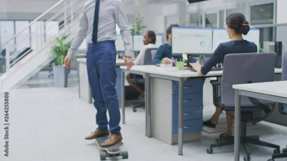 In the Bright Corporate Office Cool Businessman Wearing Tie and Shirt Rides Skateboard through Office Hallway. In the Background Professional People Working on Desktop Computers In the Bright Corporate Office Cool Businessman Wearing Tie and Shirt Rides Skateboard through Office Hallway. In the Background Professional People Working on Desktop Computers