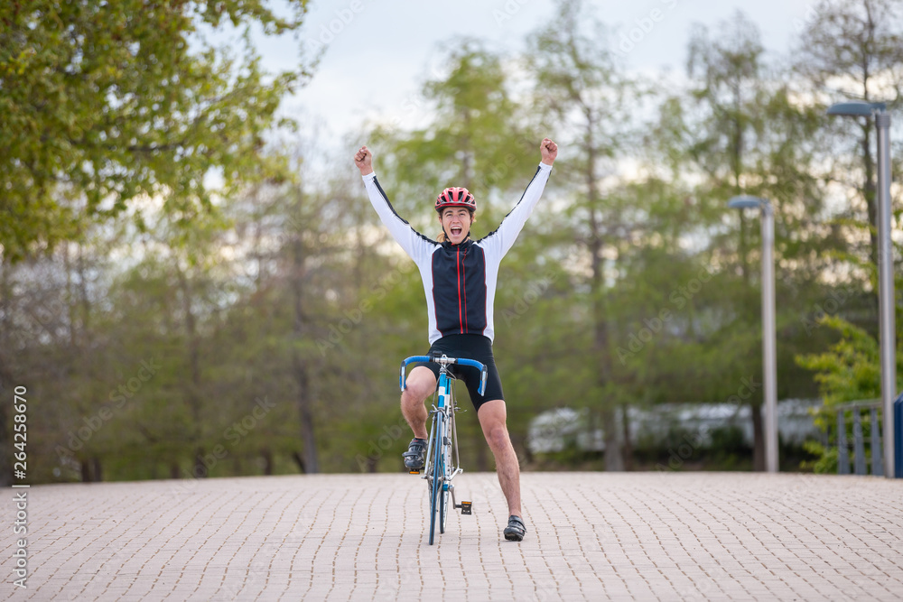 Young man in helmet and sportswear raising hands and celebrating victory while riding bicycle on pavement in park