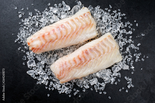 Raw Norwegian skrei cod fish filet as top view on crashed ice on a black board with copy space