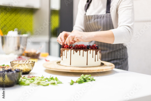 Confectioner decorates with berries a biscuit cake with white cream and chocolate. Cake stands on a wooden stand on a white table. The concept of homemade pastry, cooking cakes.