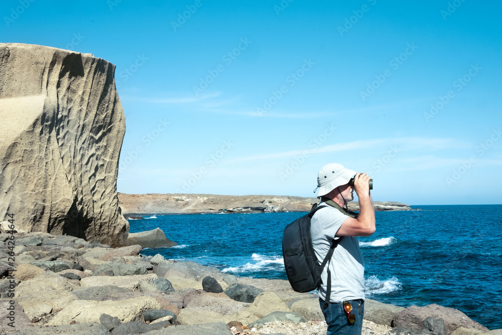 Side view of old man with white beard looking at the horizon over water with binocular. Exploring and enjoying the nature in holiday. Casual clothing