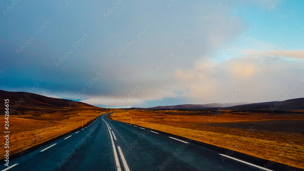An endless road through the lowlands. On the side some hills. Dark and heavy tones. Both sides of the road are barren. Sky is clouded, but still blue parts visible. Empty road, not a single car moving