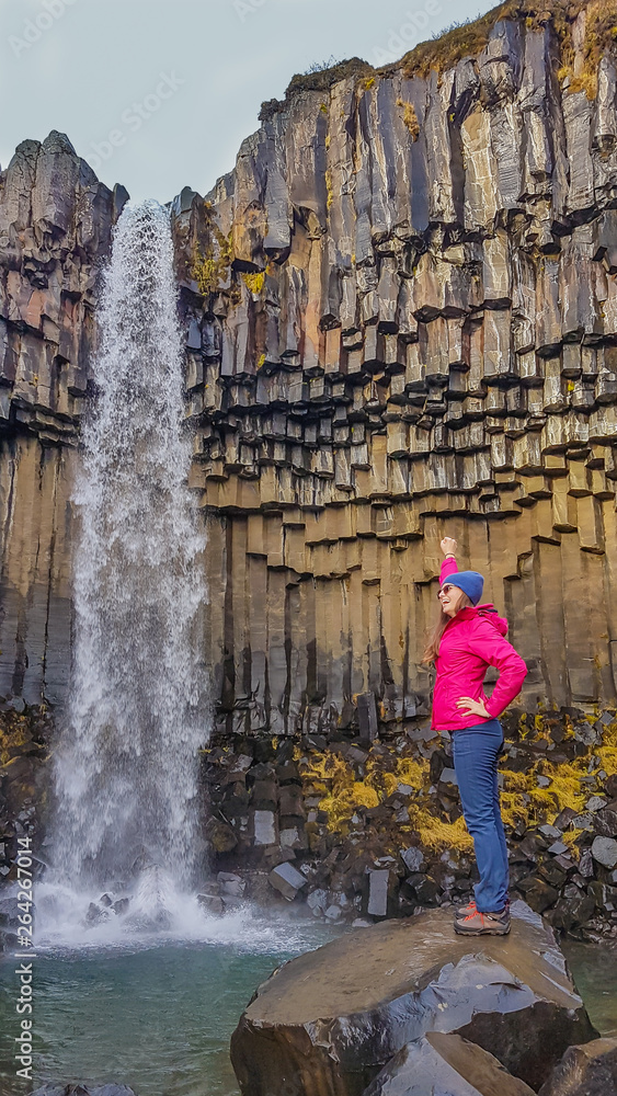 A girl wearing pink jacket, standing in front of a waterfall. She rises one hand up, powerful gesture. Rocks on the ground look slippery. The water falls from a rock formation, decorated with moss.