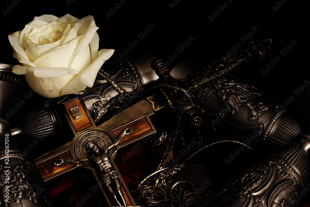Naklejka premium Stiletto, crucifix with inlaid metal chain, metal goblets for wine and white rose on black background. Souvenirs from Germany. Collecting.