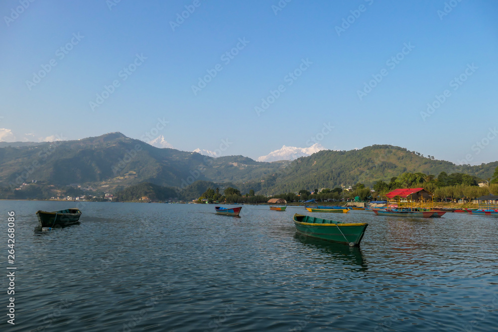 Many colourful boats parked on the shore of Phewa lake, Pokhara, Nepal. In the back Himalayan mountain range. Small villages visible on the shore. Peaceful and chilled atmosphere. Place to relax.