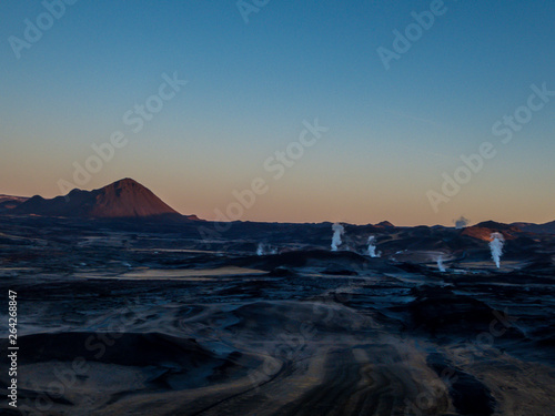 A volcanic, geothermal landscape captured during the sunrise. First beams of the sun reach the top parts of volcano. Smoke coming out of hot springs. Volcanic lava fields all around. Cosmic and alien