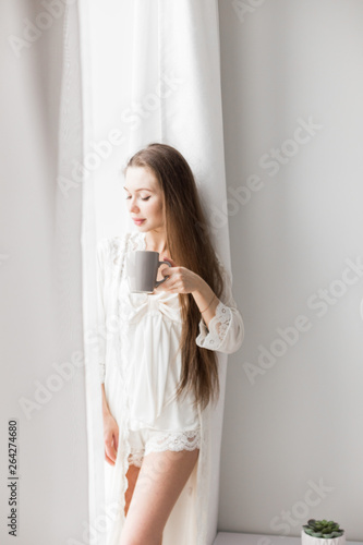 Attractive young girl in night clothes standing by the window with a mug of coffee