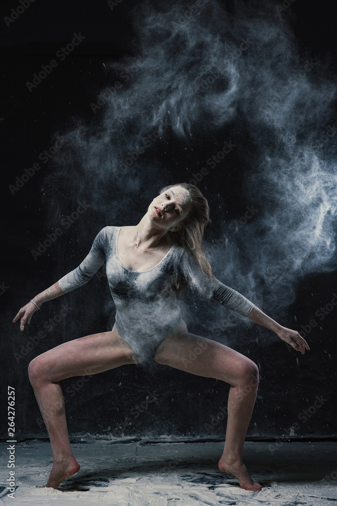 A young beautiful girl in a swimsuit and a translucent skirt emotionally dancing on a black background in clouds of white dust A young beautiful girl in a swimsuit and a translucent skirt emotionally dancing on a black background in clouds of white dust