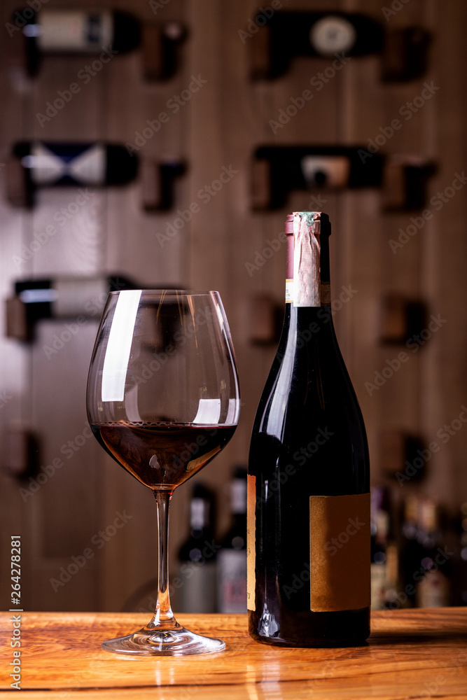 Glass and bottle with delicious red wine on wooden table. In the background Red wine bottles stacked on wooden racks shot with limited depth of field.