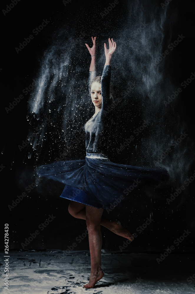 A young beautiful girl in a swimsuit and a translucent skirt emotionally dancing on a black background in clouds of white dust