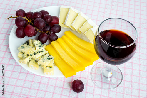 Glass of red wine and white plate with tree kinds of sliced cheese and sweet red grapes. Cheeses covered with edible white and blue mold. Food still life.