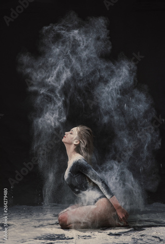 A young beautiful girl in a swimsuit and a translucent skirt emotionally dancing on a black background in clouds of white dust