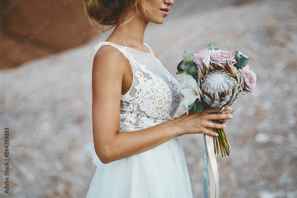 Beautiful blonde model girl with modeling wedding hairstyle in a fashionable white lace dress with a bouquet of exotics flowers in her hands