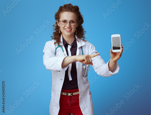 smiling elegant doctor woman pointing at smartphone on blue