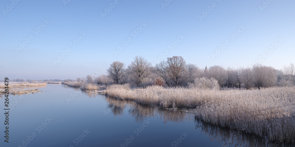 Winterlandschaft - Mit Raureif bedeckte Bäume und Schilf an Fluss vor blauem Himmel