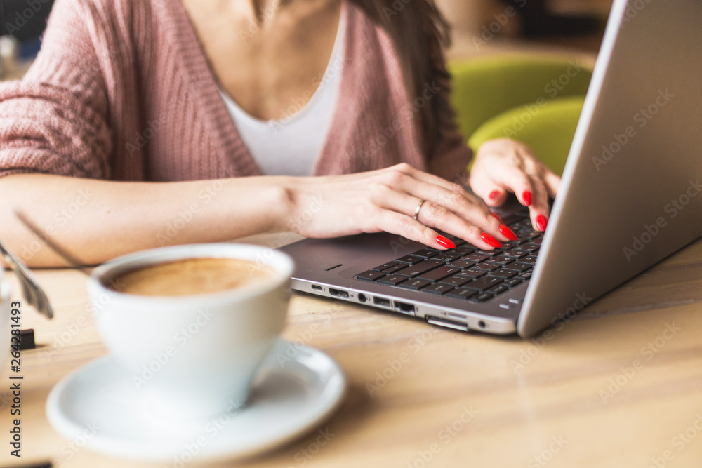 Fototapeta premium Attractive young woman sits at a table in a cafe with a cup of coffee and enjoys a laptop