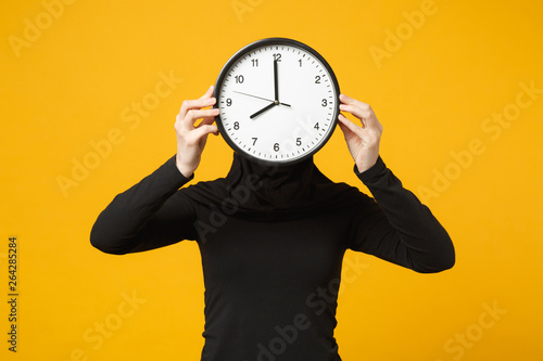 Young arabian muslim woman in hijab black clothes hold in hands round clock isolated on yellow wall background studio portrait. People religious lifestyle, time management concept. Mock up copy space.
