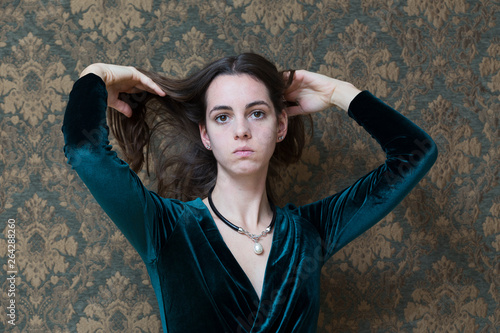 Medium horizontal shot of beautiful serious young woman in velvet dress and pearl pendant lifting her long brown hair photo