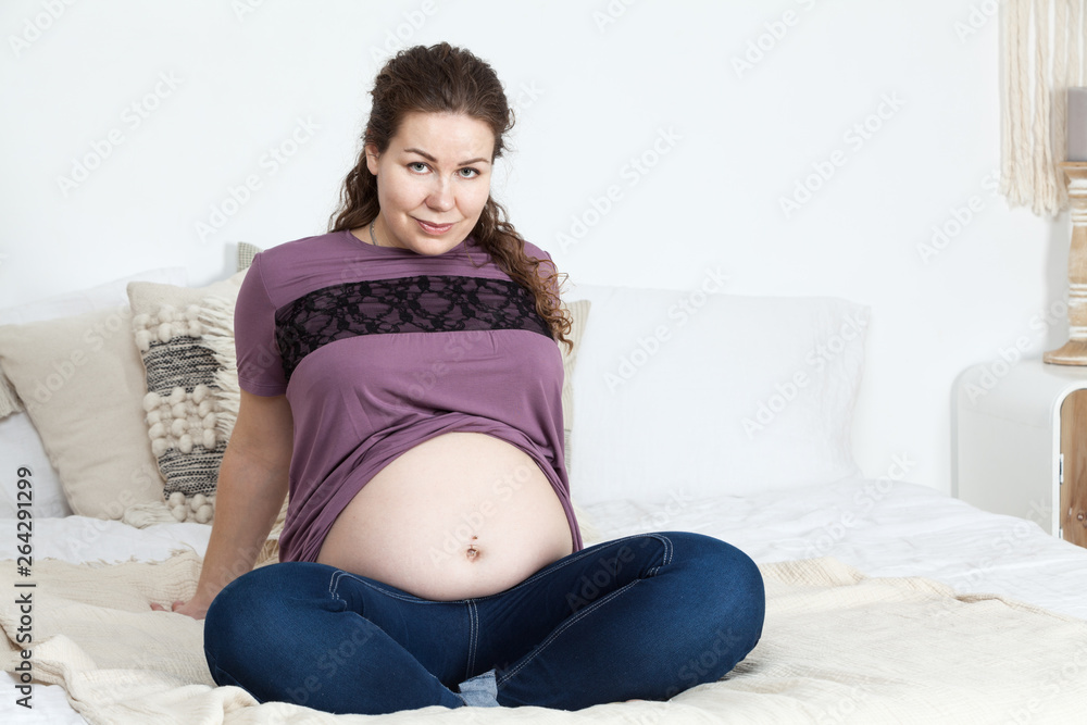 Portrait of joyful and smiling pregnant woman with naked belly, sitting on the bed in bedroom, looking at camera Portrait of joyful and smiling pregnant woman with naked belly, sitting on the bed in bedroom, looking at camera