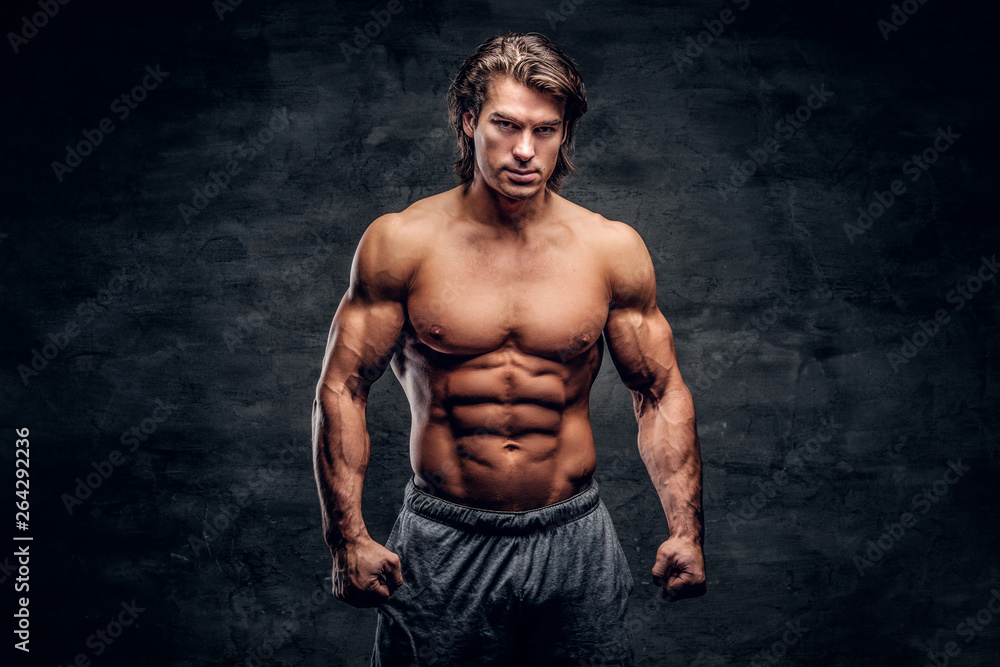 Young attractive bodybuilder with naked torso is posing for photographer on grey background.