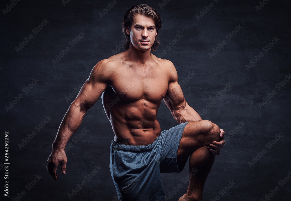 Fototapeta premium Smiling attractive bodybuilder with naked torso is doing leg strechening on grey background.