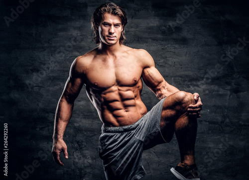 Smiling attractive bodybuilder with naked torso is doing leg strechening on grey background.