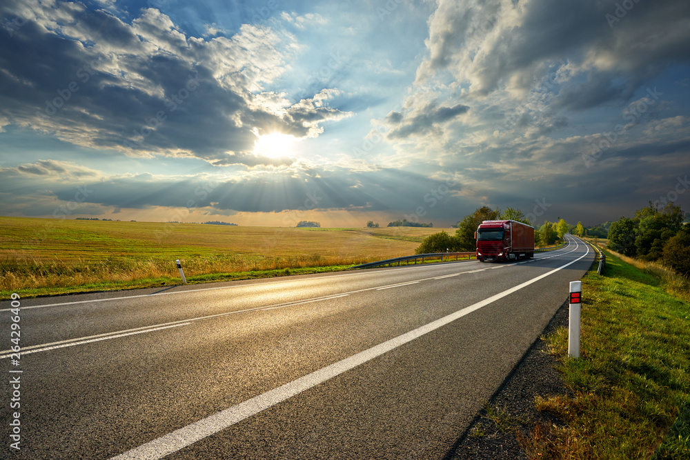 Red truck driving on the asphalt road in rural landscape at sunset with dramatic clouds