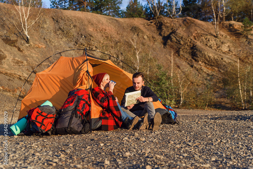 Hipster caucasian lovely couple exploring map and drinks tea or coffee in the mountain while they are sitting in the tent at sunset.