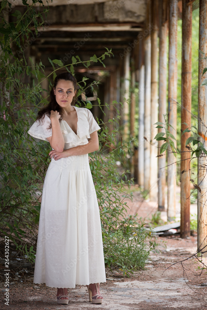 Young woman in a long white dress poses under an arcade overgrown with greenery