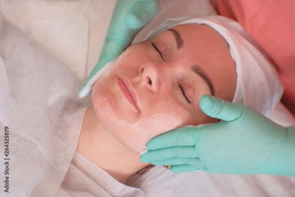Naklejka premium The girl goes through facial treatments in the beauty salon. Beautician wearing gloves apply moisturizer on the woman's face. Facials at the Spa