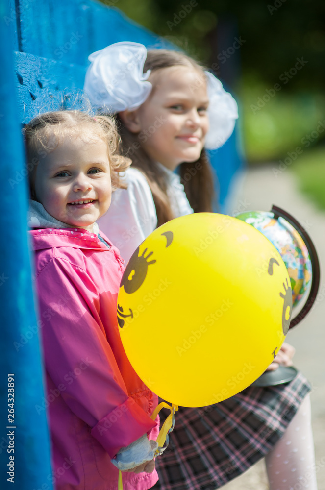 The younger sister holds a balloon and sits next to her older sister in school uniform with a globe in her hands The younger sister holds a balloon and sits next to her older sister in school uniform with a globe in her hands